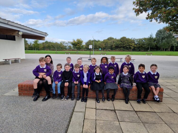 Class P1S from Heathryburn School. The children are sitting on a short brink wall outside the school in two rows, with a member of staff sitting to the left of the class