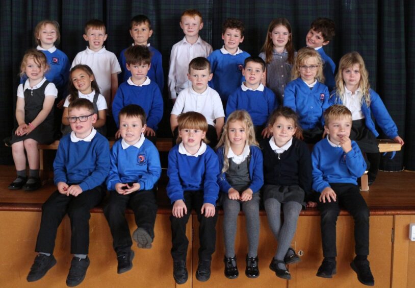 Hythehill Primary School's class P1B in three rows on the assembly hall stage with a plaid curtain behind them