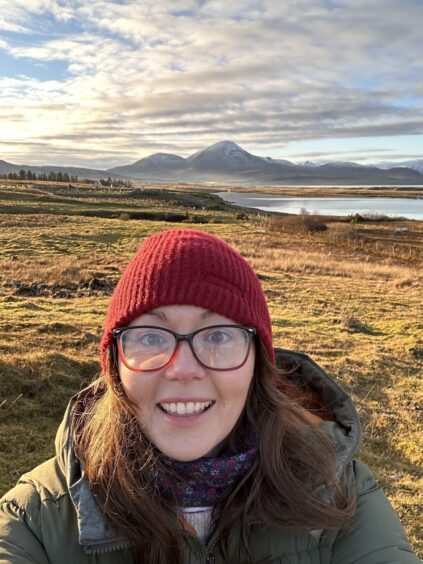 Emma Bee outside in Skye with glasses and a beanie smiling for a selfie. 