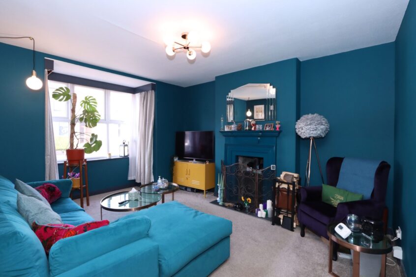 The living room, full of blue tones, including blue walls, a navy armchair, an L-shaped sofa and a fireplace. There is a large house plant next to the window, a yellow TV unit, a glass coffee table and a statement mirror above the fireplace.