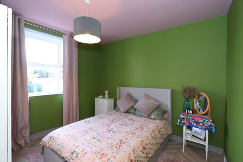 A bedroom with green painted walls, a neatly-made double bed, two white side tables and a pink rug with matching curtains