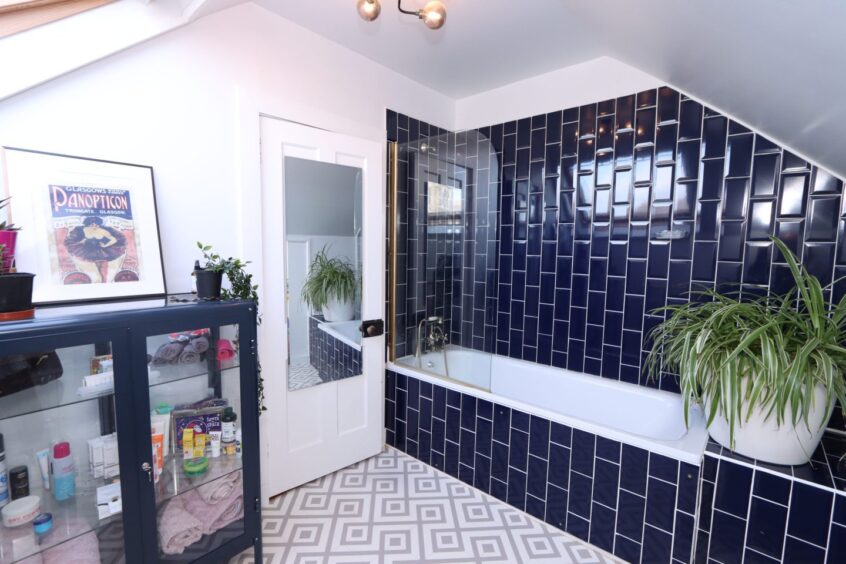 The bathroom in the renovated aberdeen home with geometric floor tiles, a glass display cabinet and a large houseplant on the end of the bath. The bath/shower has dark blue vertical tiles around it.
