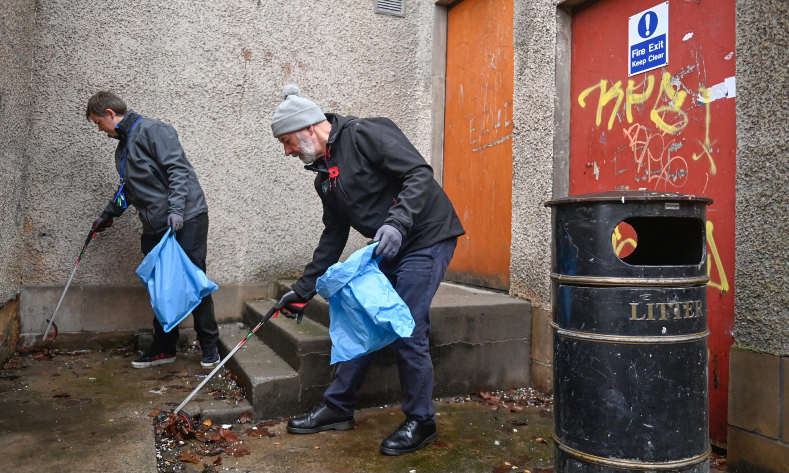 Sam and Willie picking up rubbish into blue bin bags. 