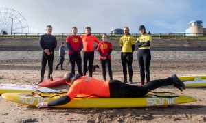 Adam Rofe giving a paddling example by lying down on a surf board on Aberdeen beach while other members watch.