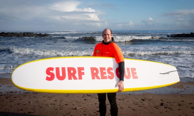 ASLSC captain Adam Rofe standing with a surf board under his arm on Aberdeen beach and smiling at the camera.