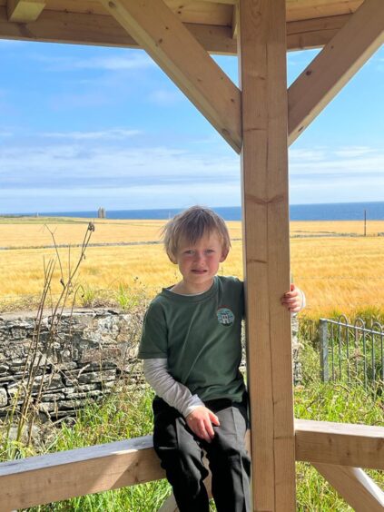 One pupil from Keiss Primary School, sitting on a wooden fence with fields behind him, the sea in the distance