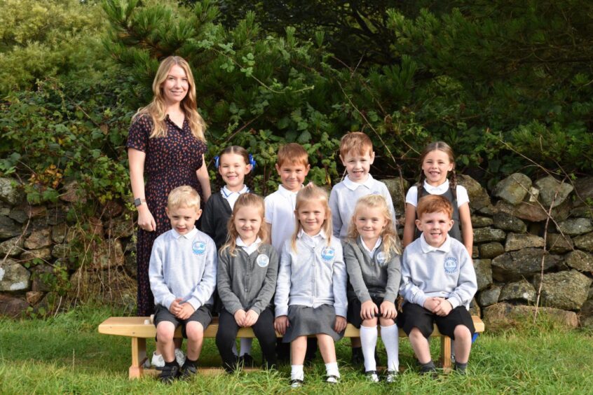 A small class sitting outside Loirston Primary School with their teacher, a short cobbled stone wall behind them along with some bushes