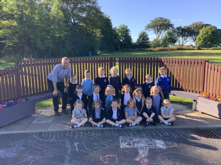 Longside Primary School's first class of 2023 in the playground, chalk drawings on the tarmac below them and a field of grass behind them. They are arranged in three rows with their teacher standing beside them.