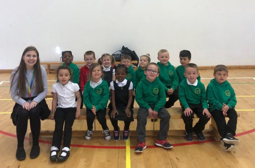 First class of 2023 at ManorPark Primary School in Aberdeen with one of their teachers sitting at the edge of the front row. The pupils are sitting on benches in two rows in the gym hall of the school