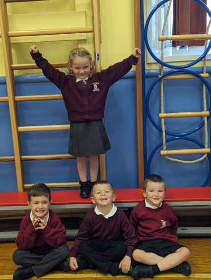 Four pupils at Maud Primary School, three boys sat on the floor and one girl standing on a bench with her arms in the air