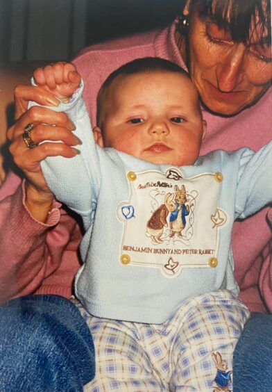 A younger Mrs Chiz shown in a pink sweatshirt holding baby grandson Ethan who is wearing tartain pyjama bottoms and a matching Peter Rabbit-themed fleece top.