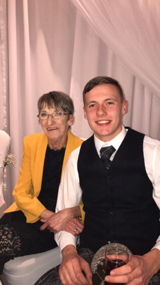 Denise Chisholm - known as Mrs Chiz - pictured with Grandson Ethan at a wedding. She is wearing a black dress and buttercup yellow jacket. Ethan is shown wearing a kilt outfit, with a black waistcoat and tartan tie, holding a drink.