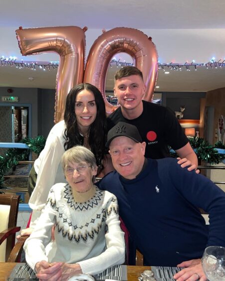 Shown in front of two metallic rose balloons that form the number 70, is Mrs Chiz - Denise Chisholm (in a Fair Isle jumper) being hugged by son-in-law Shaun Marwick, with daughter Alanna and grandson Ethan behind her. 