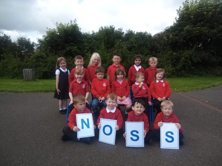 Noss Primary School's class P1.2. The children are in three rows outside the school with the front four pupils holding signs spelling out 'NOSS'