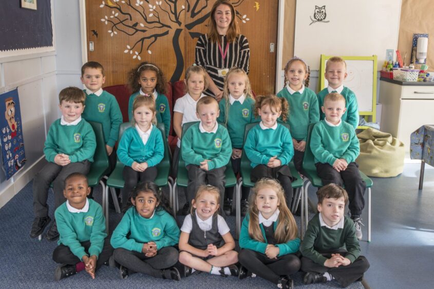 Class P1L at Riverbank Primary School in three rows of 5/6 pupils. Their teacher is standing behind them a mural of a tree on the classroom door behind them