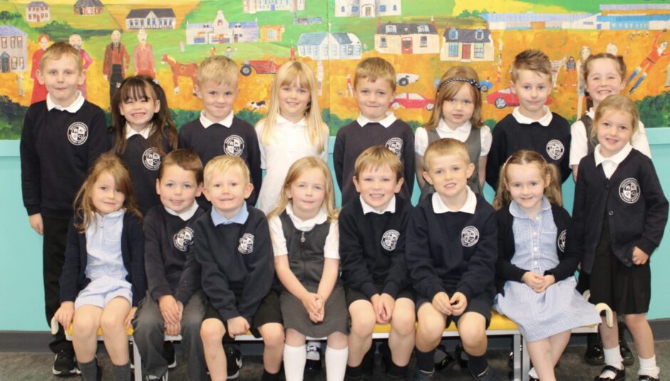 First class 2023 at Aberdeenshire school Rothienorman Primary. The pupils are in two rows in front of a wall with a colourful mural, one row standing and the other sitting on a bench at the front.