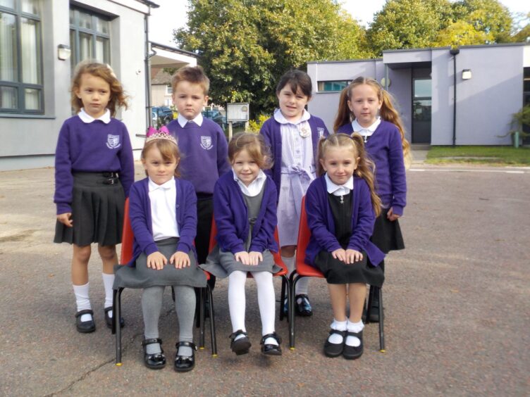 Seafield Primary School's class P1.2 in two rows, one row of four standing behind a row of three sitting on red chairs. They are pictured outside the school building