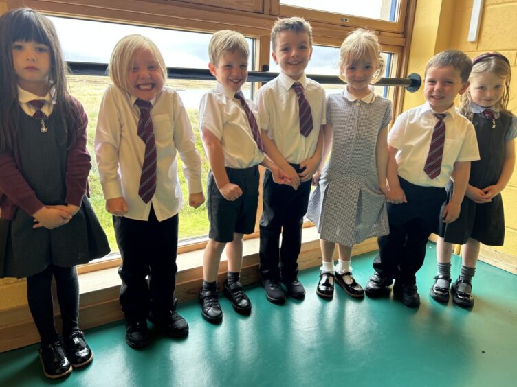 First class of 2023 at Sgoil nan Loch in the Highlands and Islands. The children are standing in a row of seven with windows overlooking the countryside behind them