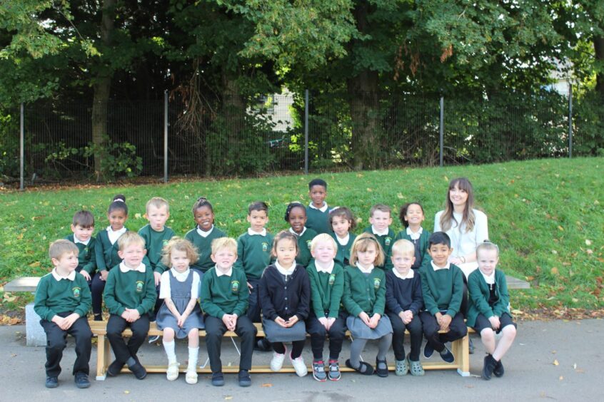 Class P1V with their teacher Miss Vivian. The pupils are in two rows outside the school with a grass hill behind them