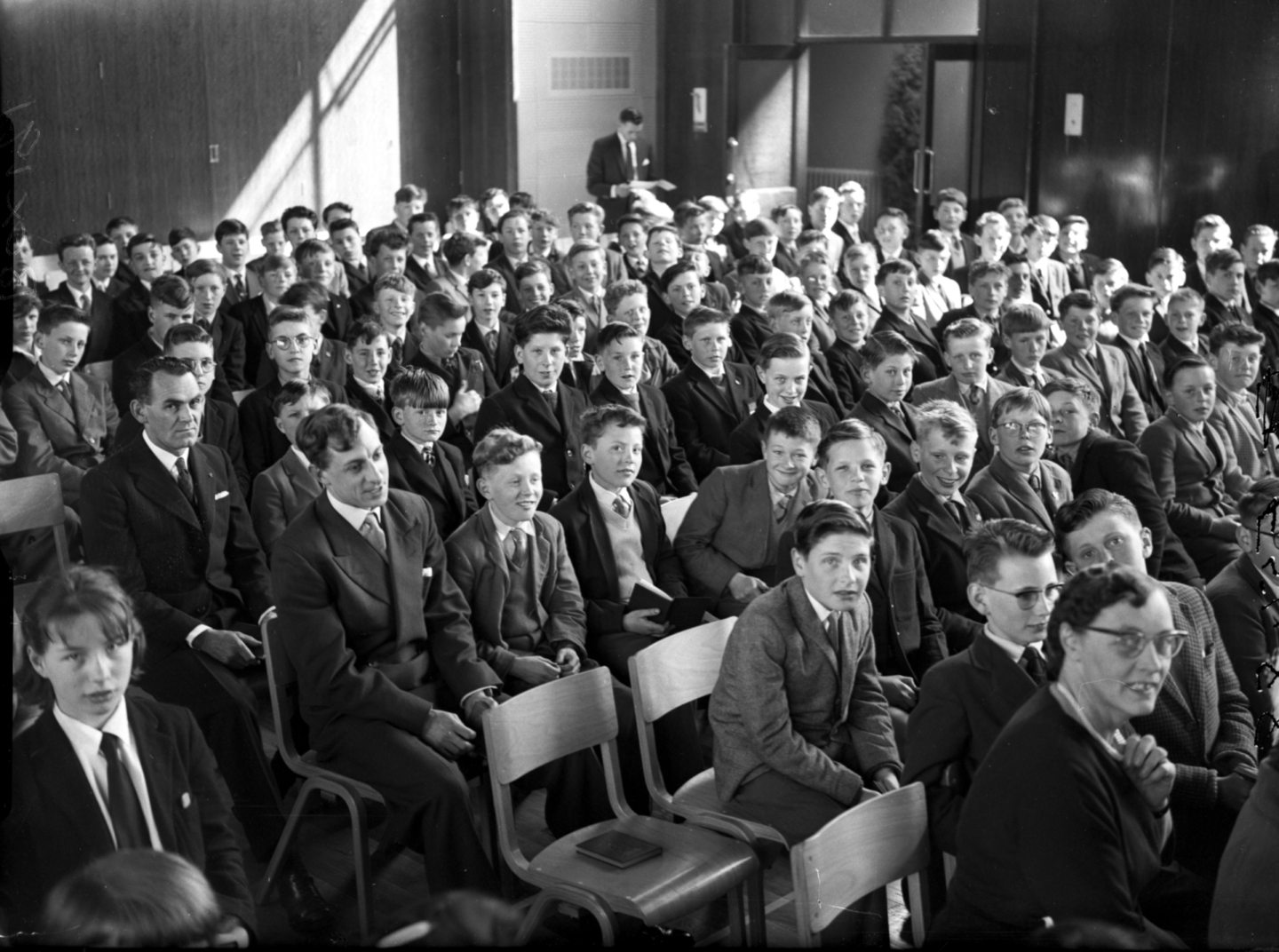 Schoolchildren and teachers sitting in neat rows on chairs in a school assembly hall look at the camera as their photo is taken.
