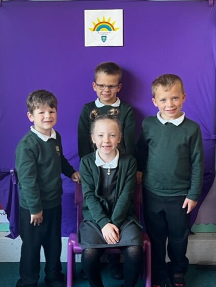 Four Sunnybank School pupils, three boys standing up and one girl sitting down on a plastic purple chair at the front. They are smiling in front of a bright purple wall, a sign with a rainbow and sun above them