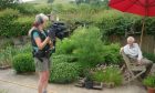 A woman with a professional film camera set-up talks to a man in a garden.