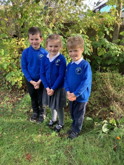 Three pupils from Tullynessle Primary School, standing outside surrounded by greenery