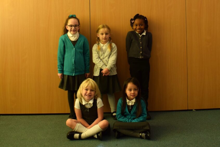 Mrs Ross's class P1-2AR, Five pupils from Walker Road School, all girls. Three of the girls are standing against a wooden wall and two are sat on the floor with their legs crossed