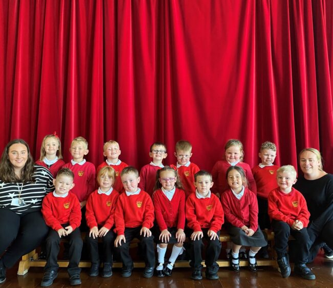 Class P1P in two rows in front of a red curtain at Westpark School with two members of staff