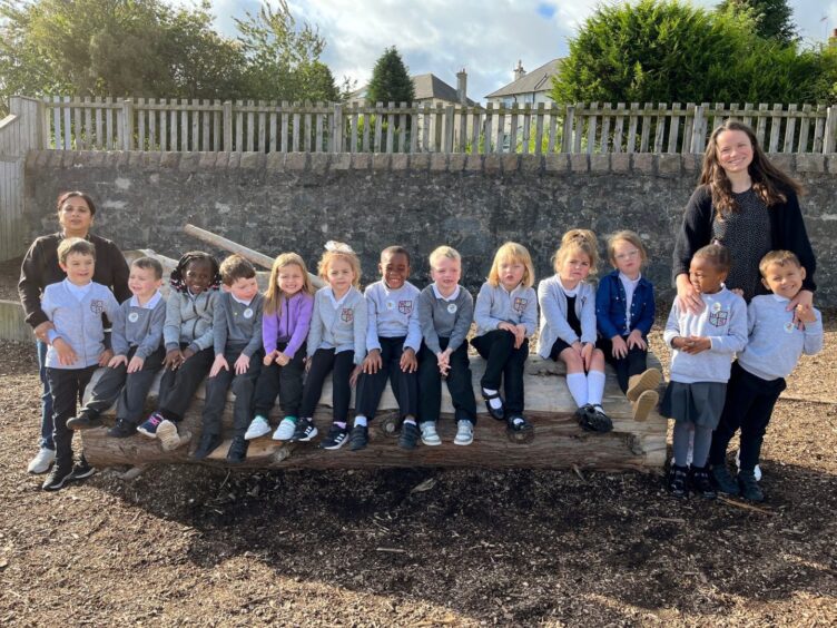 First class of 2023 at Woodside Primary in Aberdeen, the pupils are siting in a row on a rustic wooden bench with teachers on either side