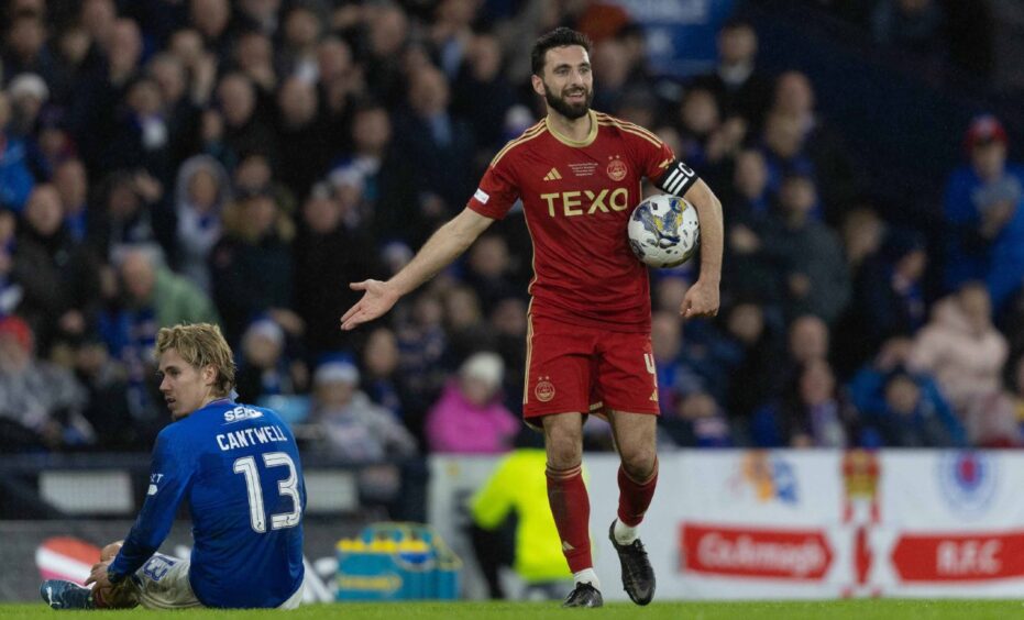 Aberdeen FC captain Graeme Shinnie standing with the ball under his arm on the pitch with Rangers' Todd Cantwell sitting on the ground behind him
