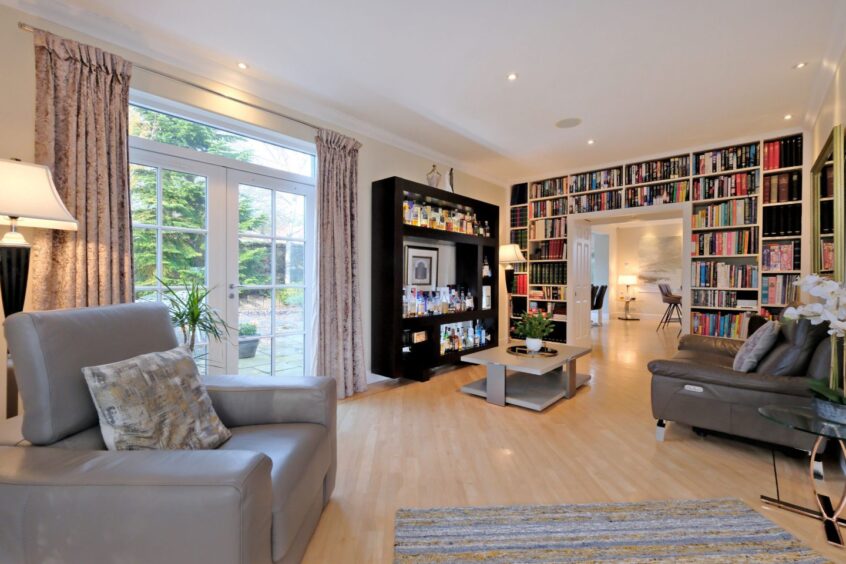 The lounge in the five-bedroom house in Bieldside, which has an armchair, sofa, coffee table and built-in book case spanning one wall around a set of double doors. There are glass double doors leading outside on another wall