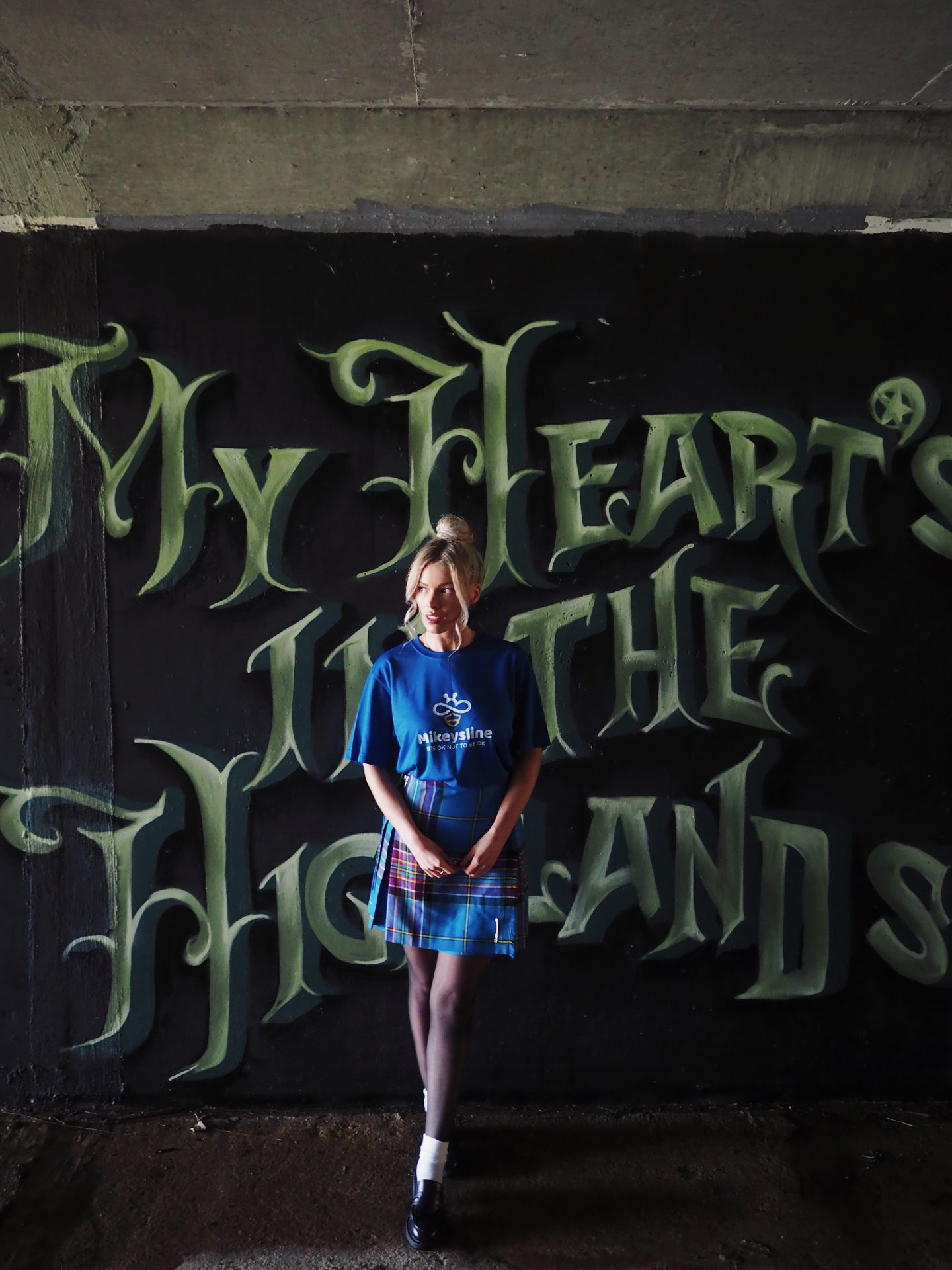 Siobhan Mackenzie wearing a Mikeysline t-shirt in front of a sign saying My heart is in the Highlands. 