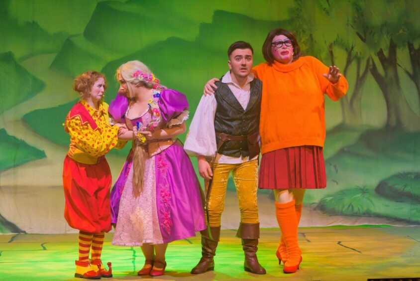 Chester, Rapunzel, Finn Flyer, and Nelly dressed as Velma from Scooby Doo wander in the woods in a scene.