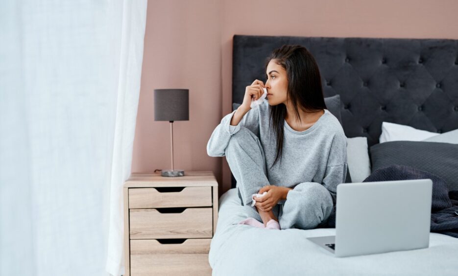 woman looks out a window and wipes her nose with a tissue while sitting on a bed with an open laptop in front of her