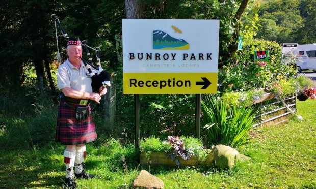 Pictured: a piper playing the bagpipes next to the welcome sign for Lochaber campsite Bunroy Park in Roy Bridge.