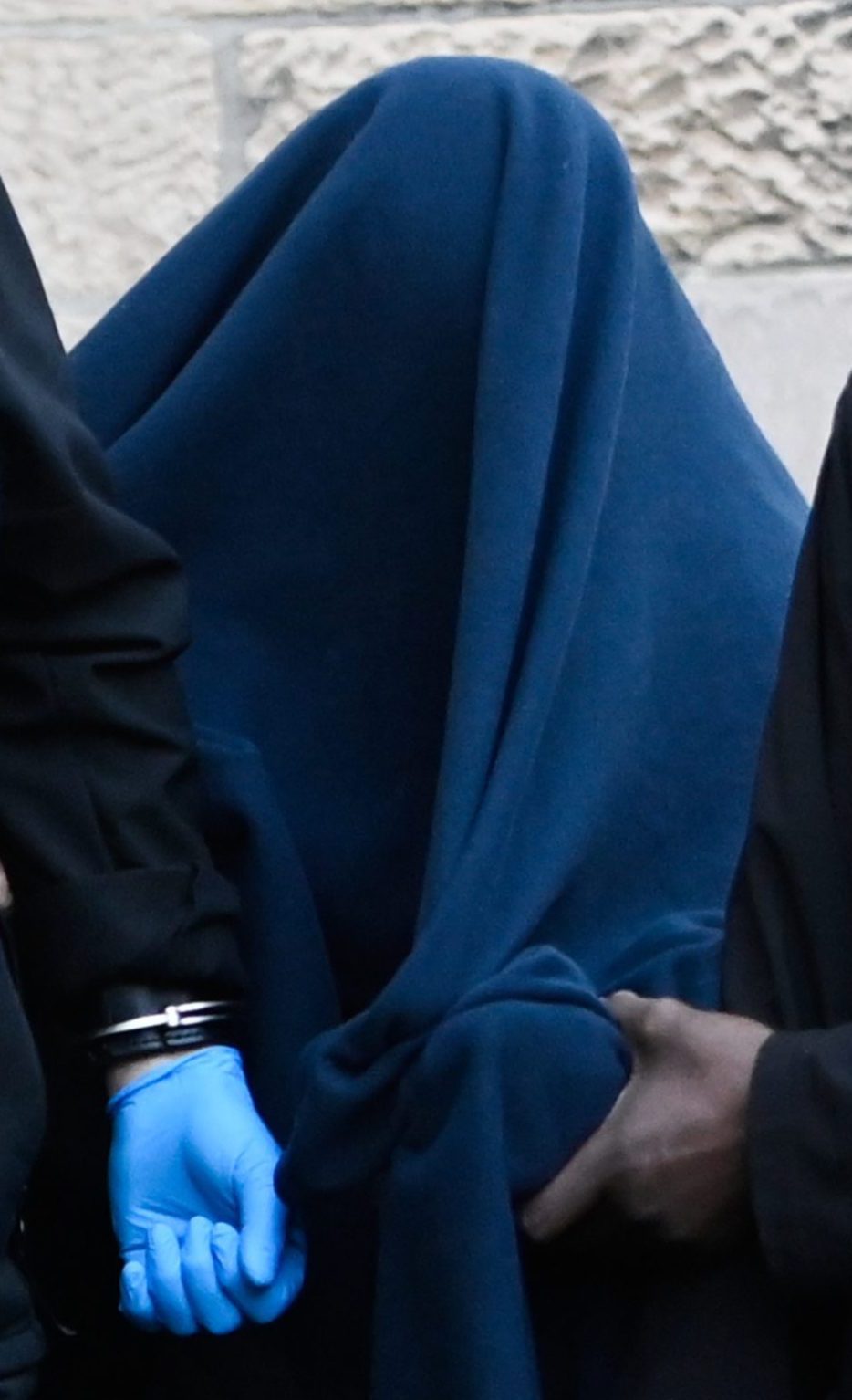 Person with blanket over their head flanked by two security guards.