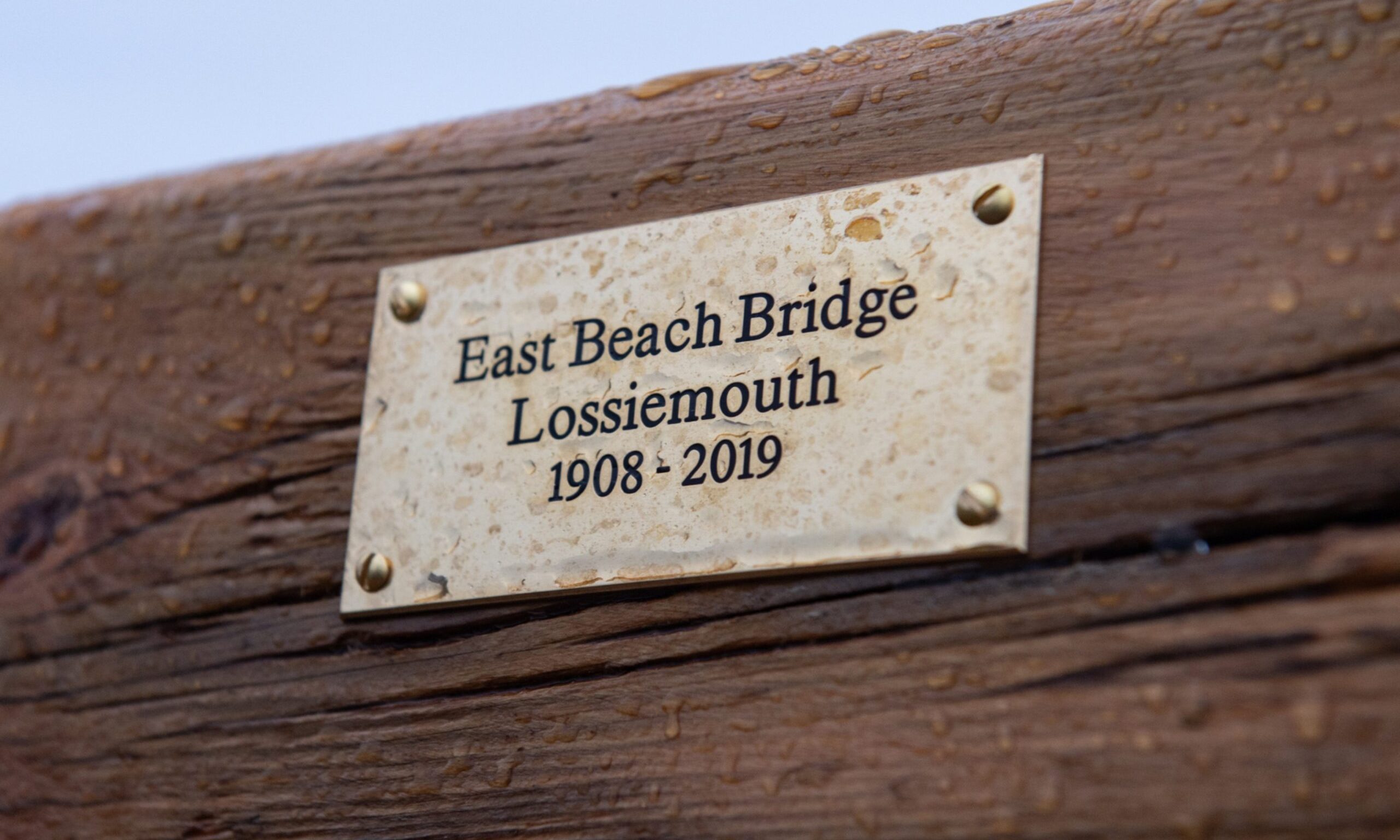 Plaque on bench reaching "East Beach Bridge Lossiemouth 1908 - 2019". 