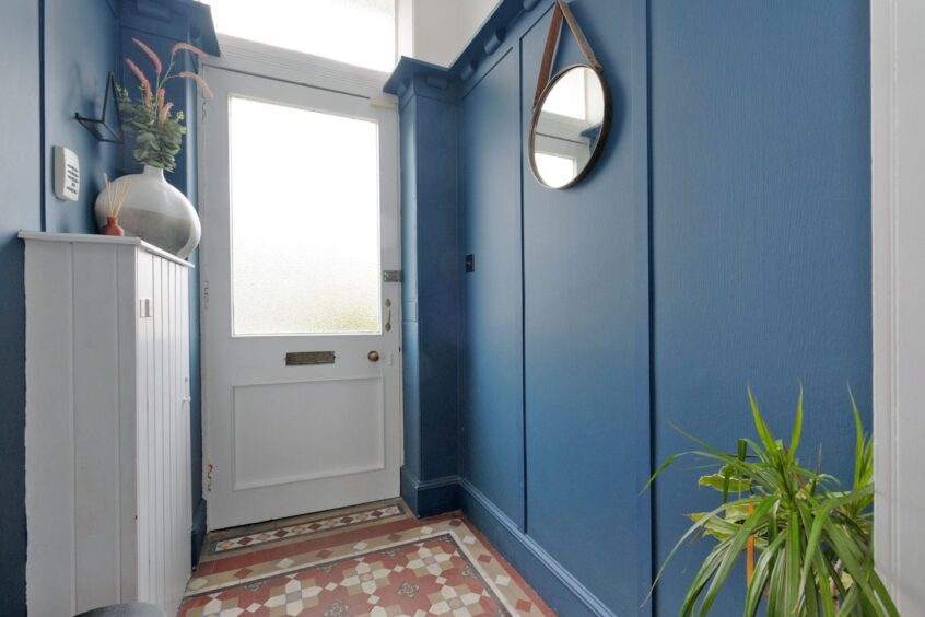The hallway to the back door has blue walls and a mosaic tiled floor. There are a couple of house plants and a round wall mounted mirror