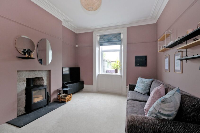 The snug of the traditional aberdeen home has a woodburning fireplace, mauve pink walls, a large dark mauve couch, two mismatching mirrors above the fireplace and a TV in the corner of the room. There are shelves on the wall above the sofa