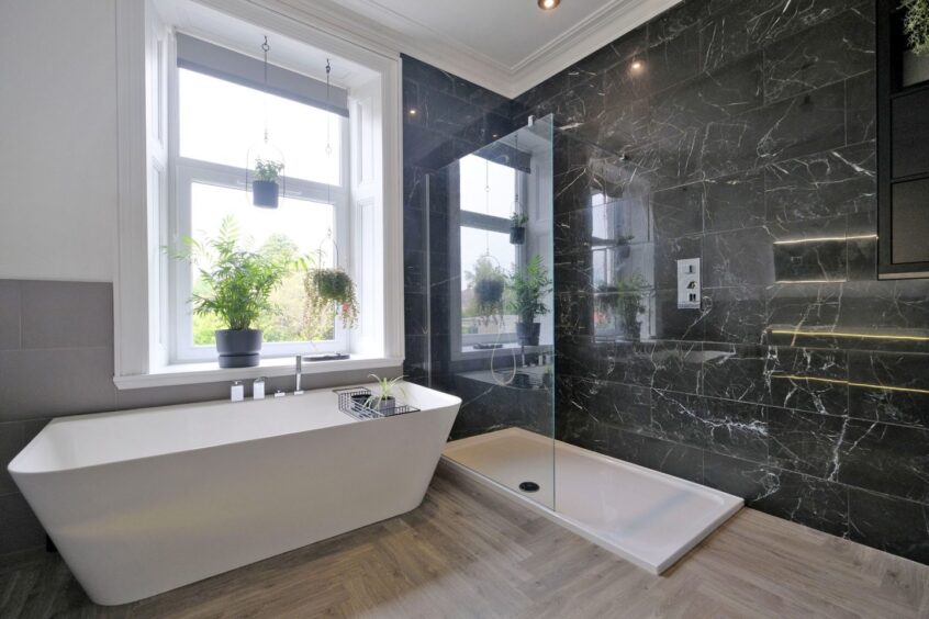 The sleek bathroom in the property with a large window above the bath, houseplants on the windowsill, a walk-in shower with a glass screen and black marble walls