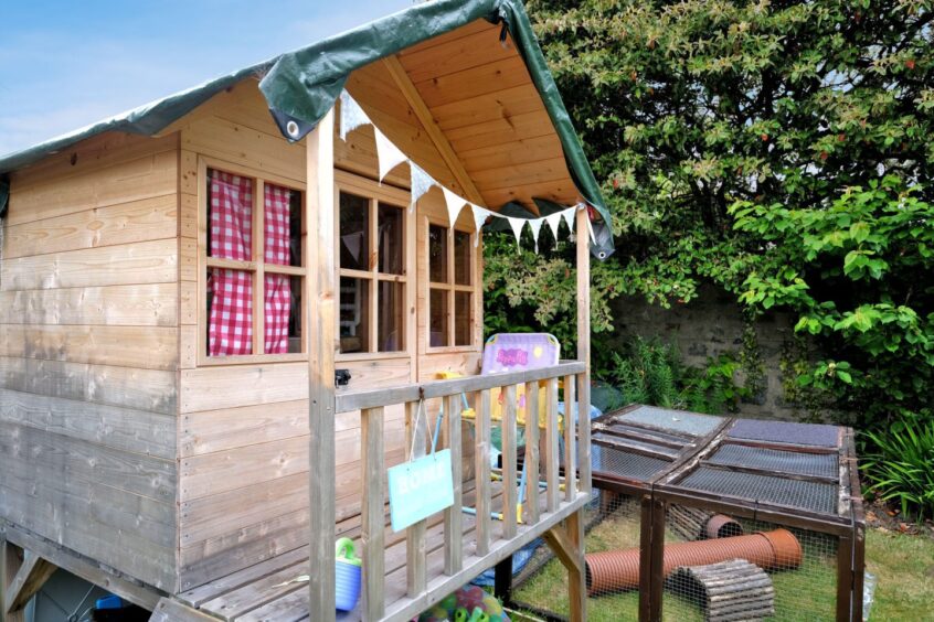 A wooden summer house, decorated with colourful bunting, gingham curtains and a colourful children's chair on the porch