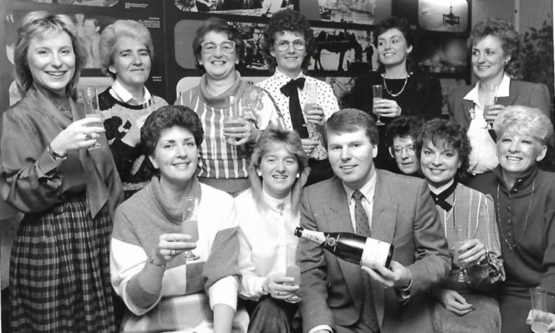 A black and white photo of women holding glasses in the air while a man pours a glass of champagne, smiling at the camera