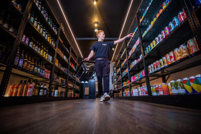 Manager Matthew Gaughan at Discovery Beers, walking down an aisle with shelves full of Scottish beer. 