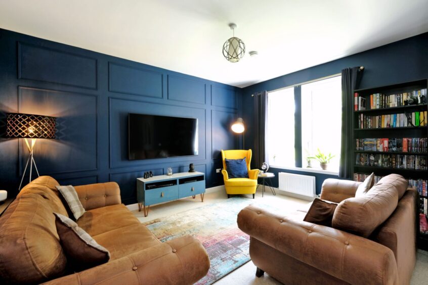 The living room in the Cove home in aberdeen has a dark blue walls, two brown leather sofas, a bright yellow armchair, a bookcase, a wall-mounted tv and a blue unit