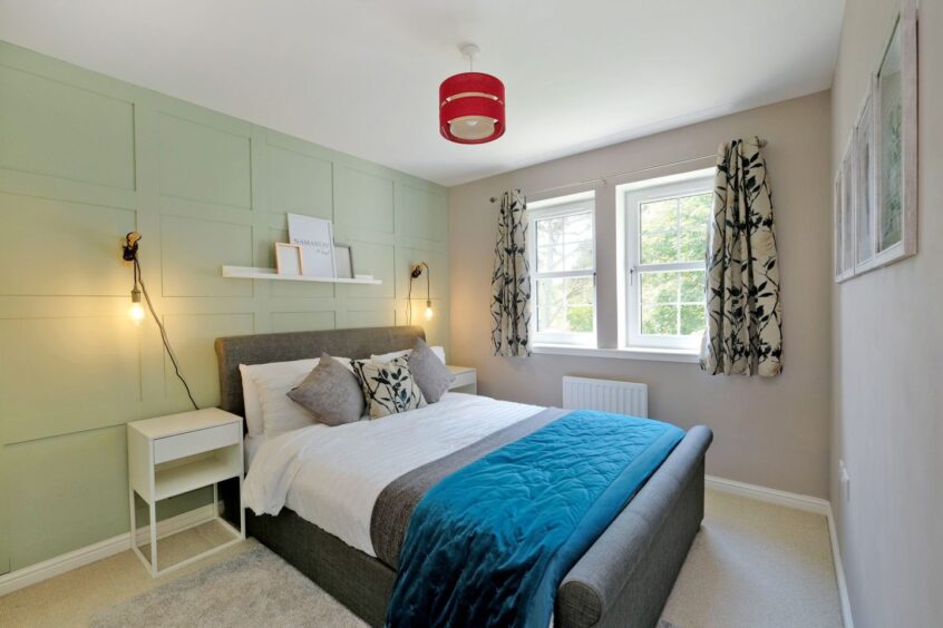A bedroom in the cove home in Aberdeen with a light green feature wall with square panels, two hanging wall lights on either side of a double bed and a shelf above the bedframe