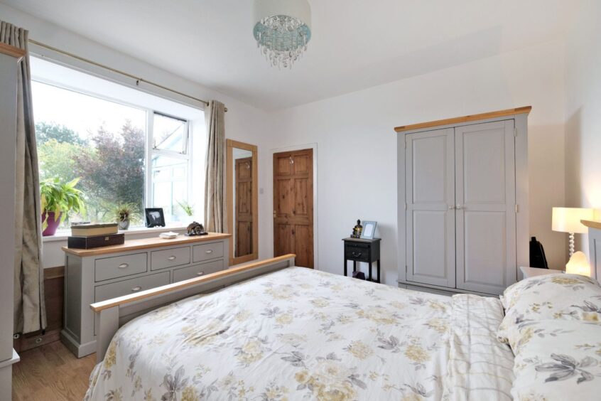 A bedroom in the country home near Aberdeen, with matching grey furnishings with wood accents. There is a wardrobe, unit of drawers, wall mounted mirror and bed.