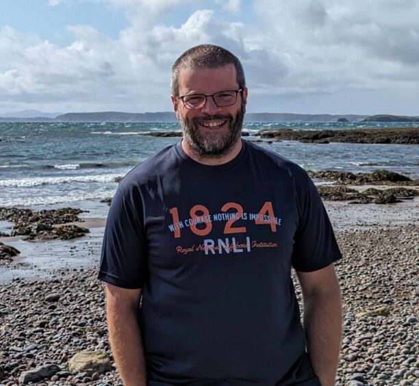 A smiling man stands in front of the sea, wearing an RNLI-branded t-shirt.