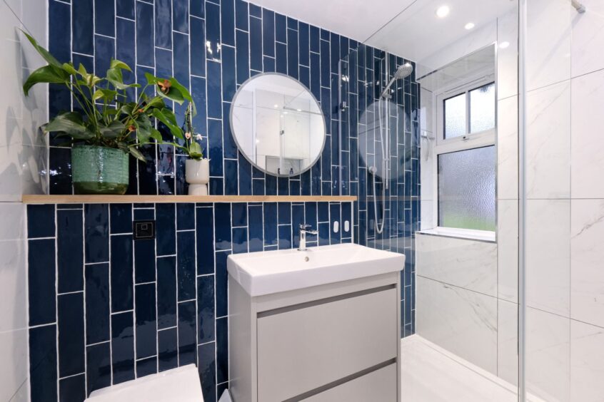 The bathroom with marble tiled walls, a navy tiled accent wall and shelf with plants on it