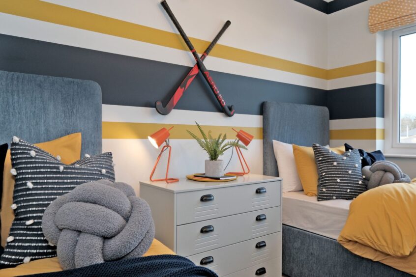 A bedroom with two single beds, navy and mustard bedding and stripes on the wall. There is a metal unit in between the beds with two bedside lamps and a plant, above it is two hockey sticks mounted on the wall